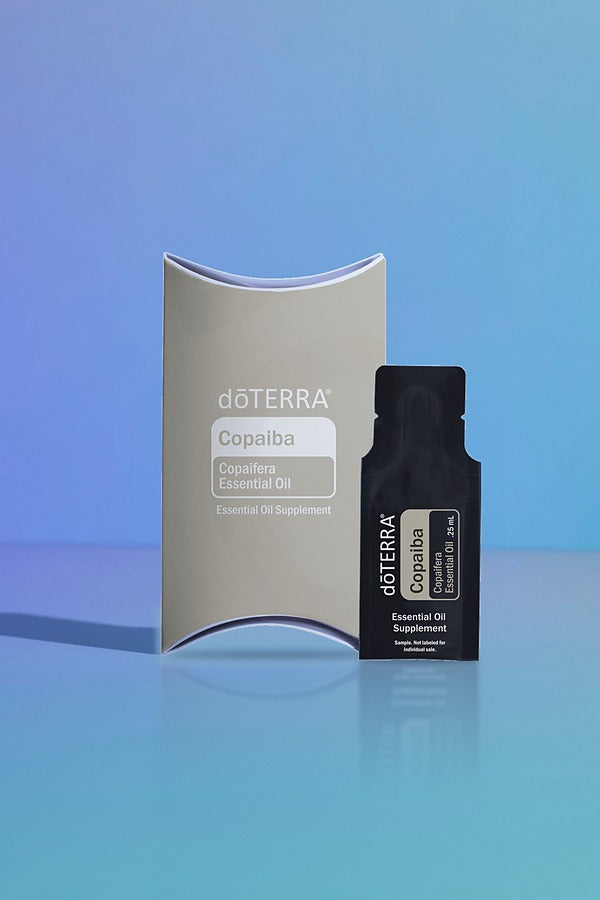 doTERRA Copaiba .25 mL black sample sachet standing beside its taupe doTERRA pillow box on a glossy blue studio surface with soft reflections. doTERRA Copaiba Essential Oil Sample (.25 mL)