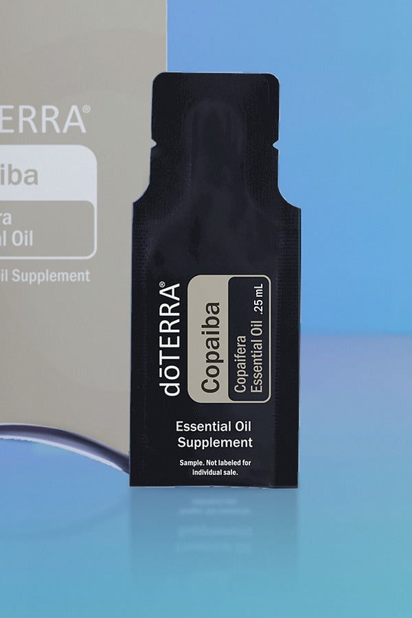 Close-up of the doTERRA Copaiba .25 mL black sample sachet standing upright in front of its taupe pillow box on a blue background. doTERRA Copaiba Essential Oil Sample (.25 mL)