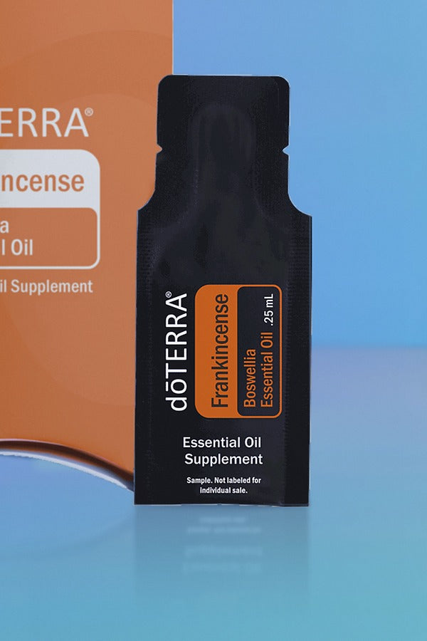 Close-up of the doTERRA Frankincense .25 mL black sample sachet standing upright in front of its orange pillow box on a blue background. doTERRA Frankincense Essential Oil Sample (.25 mL)