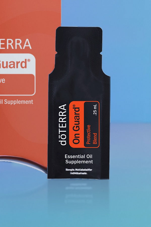 Close-up of the doTERRA On Guard Protective Blend .25 mL black sample sachet standing upright in front of its red-orange pillow box on a blue background. doTERRA On Guard Essential Oil Sample (.25 mL)