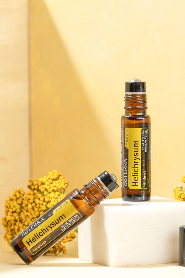 Two amber glass roll-on bottles of doTERRA Helichrysum essential oil, one standing upright on a white block and one lying diagonally, with yellow flowers against a warm beige background.