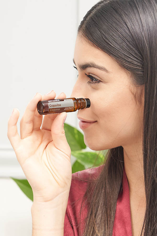 A woman holds a small amber glass bottle near her nose, showing the label which reads 'Sopapila' and 'COPPER'.