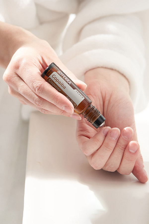 A person's hands holding a small amber roll-on bottle labeled 'DROPSA Copaliba' over their palm, with a soft white robe in the background.