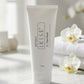 A 100g white squeeze tube of Goterra I Kise 2 Face Wash standing on a marble surface, with soft-focus white orchids and folded towels in the background.