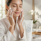 A woman in a white robe washes her face with foaming facial cleanser, with a white tube of KISE 21 Facial Wash standing on a marble countertop in the foreground.