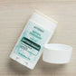 A doTERRA Natural Deodorant stick with its cap removed, lying on a light wood grain surface. The label features a white and light blue design with green text and accents.
