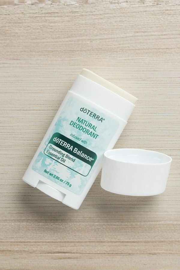 A doTERRA Natural Deodorant stick with its cap removed, lying on a light wood grain surface. The label features a white and light blue design with green text and accents.
