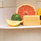 A rectangular bar of doTERRA soap in a pale yellow color, displayed on a white shelf alongside citrus fruits (grapefruit, lime, lemon) against a tiled background.