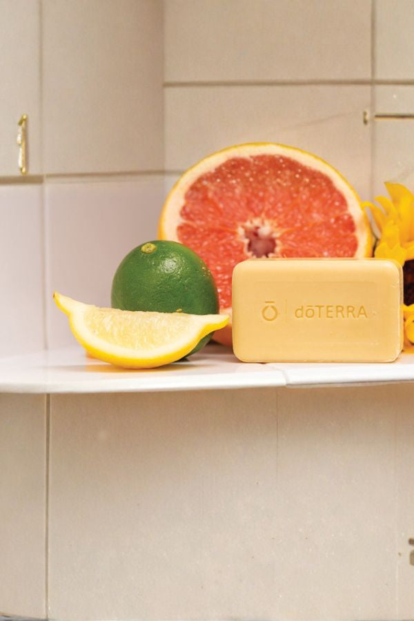 A rectangular bar of doTERRA soap in a pale yellow color, displayed on a white shelf alongside citrus fruits (grapefruit, lime, lemon) against a tiled background.