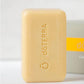 A bar of doTERRA soap in a pale yellow color, standing upright in front of its gray and yellow packaging box on a clean white surface.