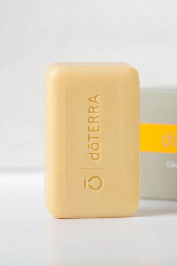 A bar of doTERRA soap in a pale yellow color, standing upright in front of its gray and yellow packaging box on a clean white surface.