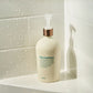 A bottle of Daily Conditioner shampoo with a white body, metallic rose gold pump, and green text, set on a white tiled bathroom ledge with water droplets.