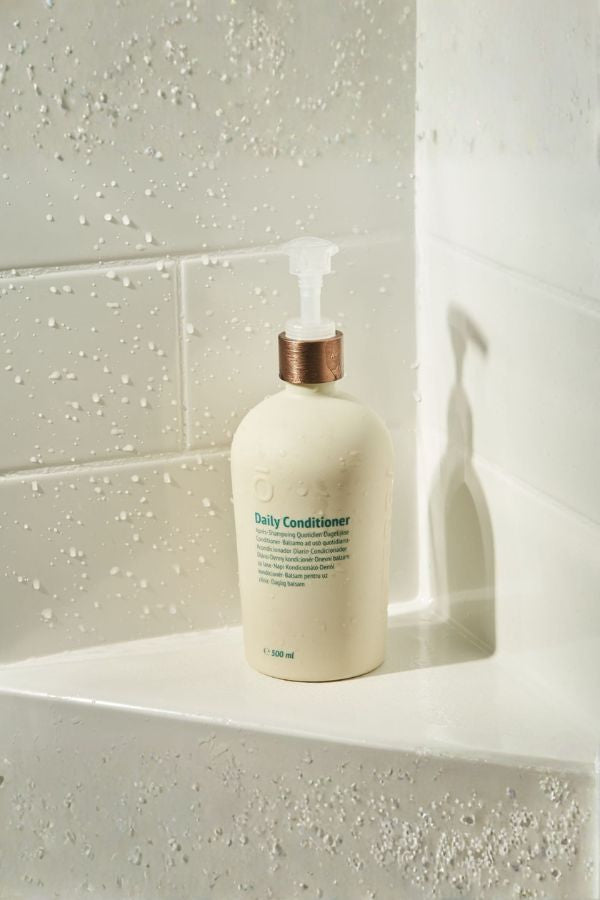A bottle of Daily Conditioner shampoo with a white body, metallic rose gold pump, and green text, set on a white tiled bathroom ledge with water droplets.