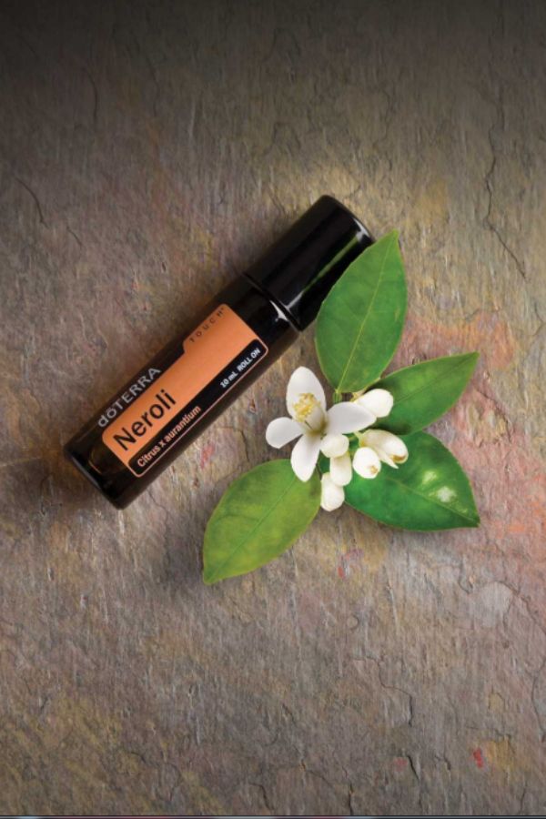 A small black dōTERRA Neroli essential oil roller bottle placed diagonally on a textured gray stone surface, styled with green leaves and white neroli blossoms.