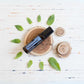 A small black roll-on bottle labeled Peppermint and d-Terpinolol, placed on a round wood slice with green mint leaves around it on a white distressed wooden surface.