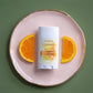 A deodorant stick package sits centered on a pale pink plate with gold trim, flanked by two orange slices, against a muted green background. The package is labeled osterra and reads Orange.