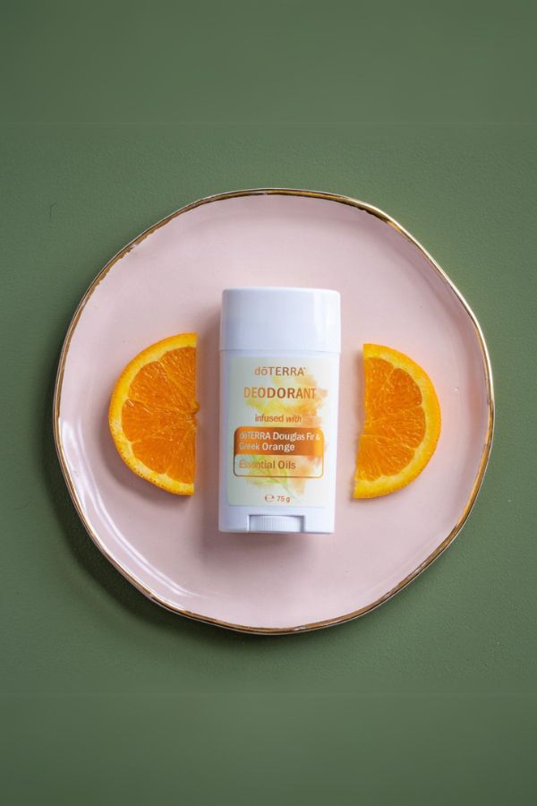 A deodorant stick package sits centered on a pale pink plate with gold trim, flanked by two orange slices, against a muted green background. The package is labeled osterra and reads Orange.