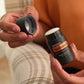 A person holds an open black doTERRA Frankincense stick hair balm jar next to its cap, with plaid beige and white trousers and an orange top visible in the background.