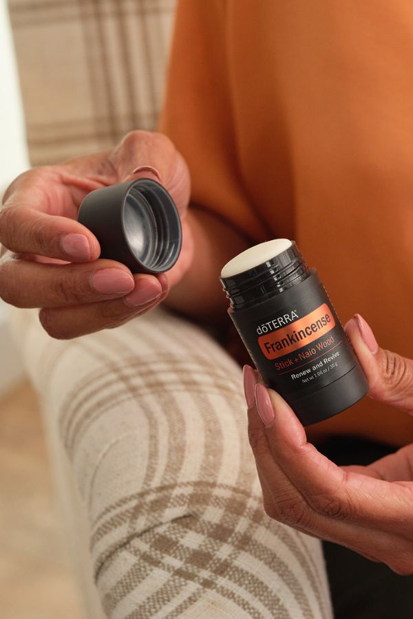 A person holds an open black doTERRA Frankincense stick hair balm jar next to its cap, with plaid beige and white trousers and an orange top visible in the background.
