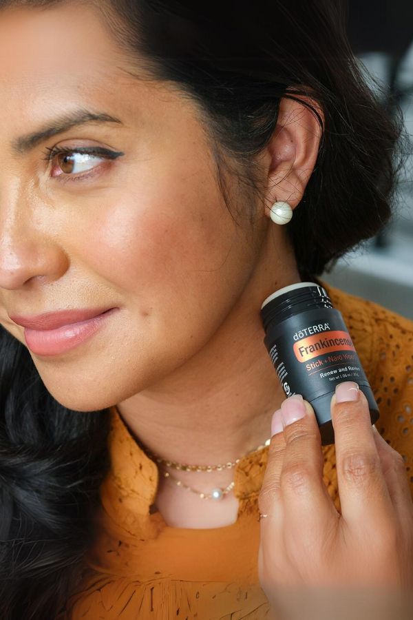 A woman holding a black Osterixx FaceMinimizing Deodorant stick next to her ear, wearing a textured orange top.
