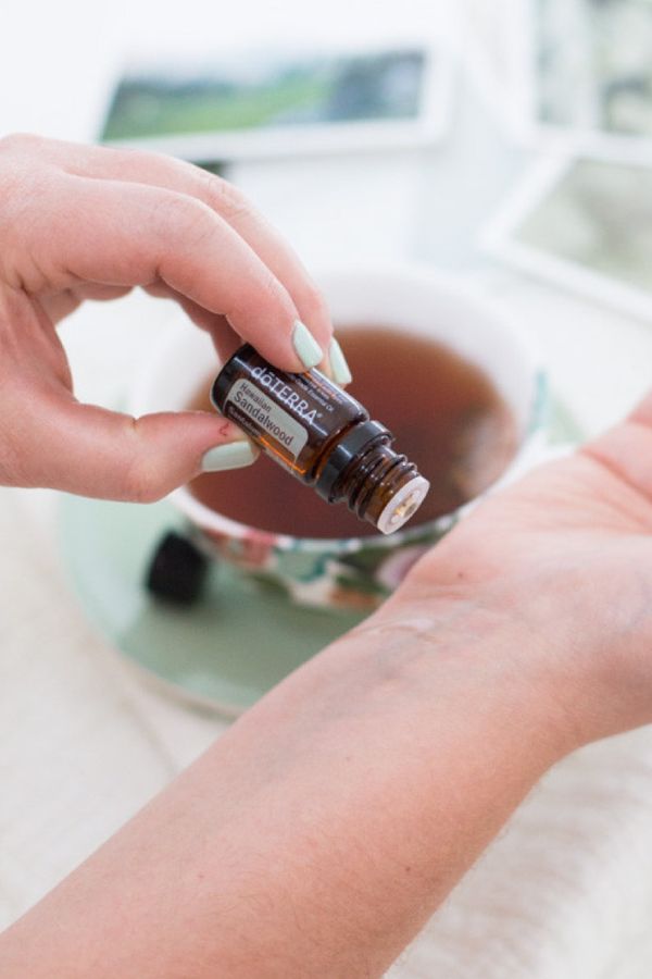 A person's hand holds a small amber glass essential oil bottle labeled 'MELINDA' above an open cup of tea, with a blurred light-colored background. doTERRA Sandalwood (Hawaiian Iliahi) Essential Oil