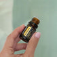 A hand with pink manicured nails holds a small amber glass bottle of doTERRA Radiocream against a soft, pale green background. doTERRA Helichrysum Essential Oil