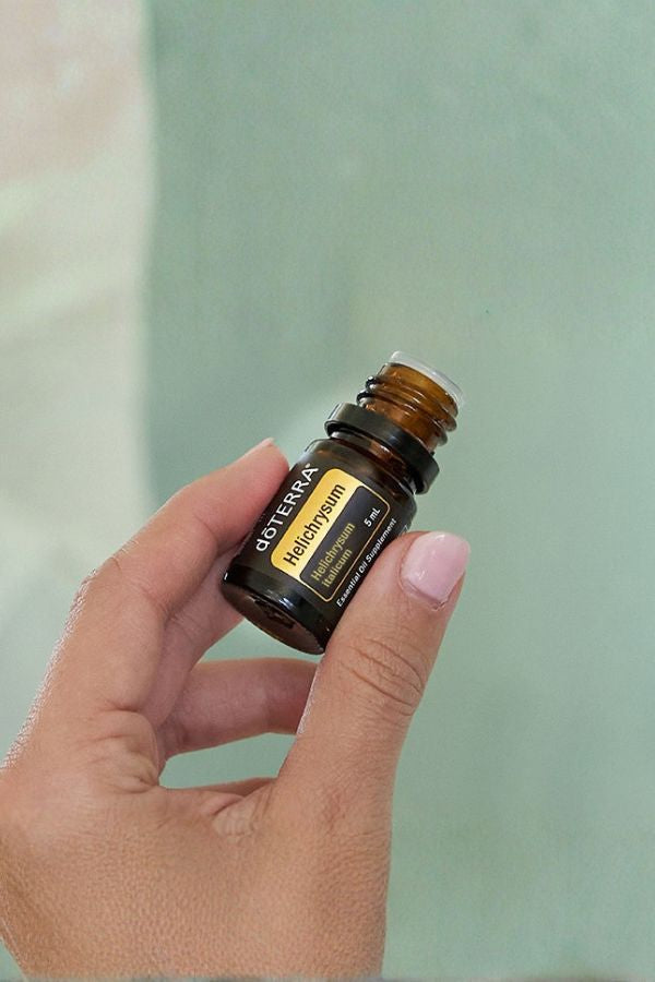 A hand with pink manicured nails holds a small amber glass bottle of doTERRA Radiocream against a soft, pale green background. doTERRA Helichrysum Essential Oil