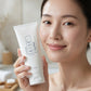 A woman holding a white tube of KISE Moisturize 2 Face Wash in front of her face, with a softly lit bathroom background.