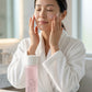 A woman in a white bathrobe applies a pink skincare cream to her face, with a pink bottle of KISE skincare resting on the marble counter in the foreground.