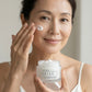 A woman applies a doTERRA IKISE Day & Night Creamto her cheek while holding a jar of KISE 2 Hour Lock Moisturizer in her other hand, set against a soft, neutral background.