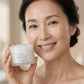 A smiling woman holds up a white jar of IKISE 11 Cleansing Balm against a soft, warm beige background.