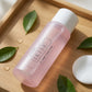 A bottle of IKISE outdoor facial cleanser with a white cap and translucent pink liquid, styled on a wooden tray with green leaves, water droplets, and a cotton pad.