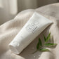 A white tube of UKISE 3-in-1 facial wash lies diagonally on a beige linen surface with water droplets and a small green leaf sprig nearby, in soft natural light.