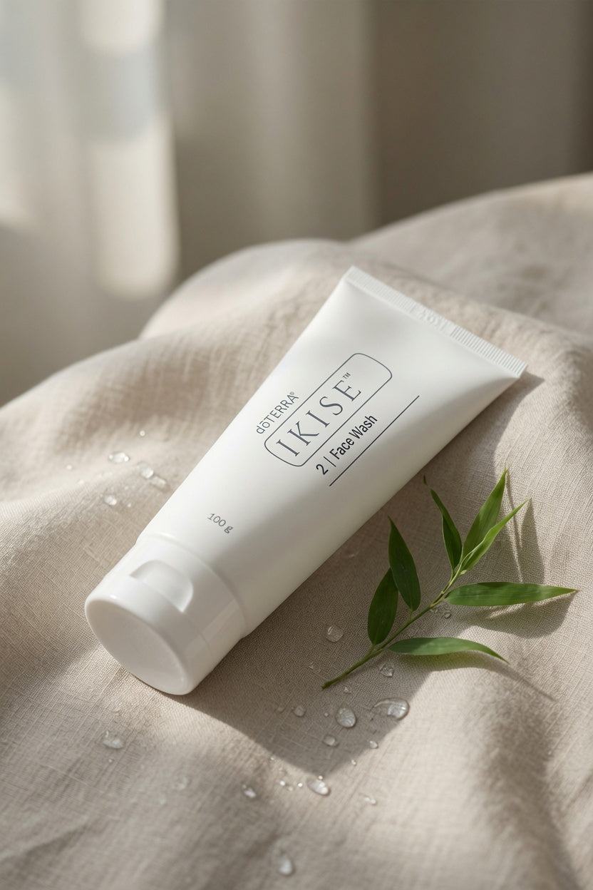 A white tube of UKISE 3-in-1 facial wash lies diagonally on a beige linen surface with water droplets and a small green leaf sprig nearby, in soft natural light.