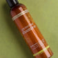 A bottle of Bionica Salon Essentials Protecting Shampoo in a translucent amber-brown bottle with a black cap, placed diagonally on a textured olive green background. doTERRA Salon Essentials Shampoo and Conditioner