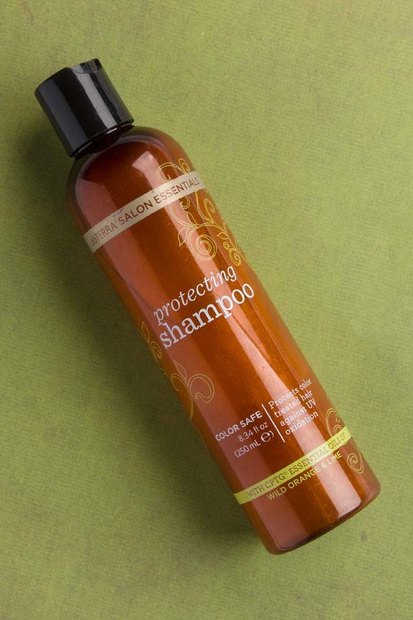 A bottle of Bionica Salon Essentials Protecting Shampoo in a translucent amber-brown bottle with a black cap, placed diagonally on a textured olive green background. doTERRA Salon Essentials Shampoo and Conditioner