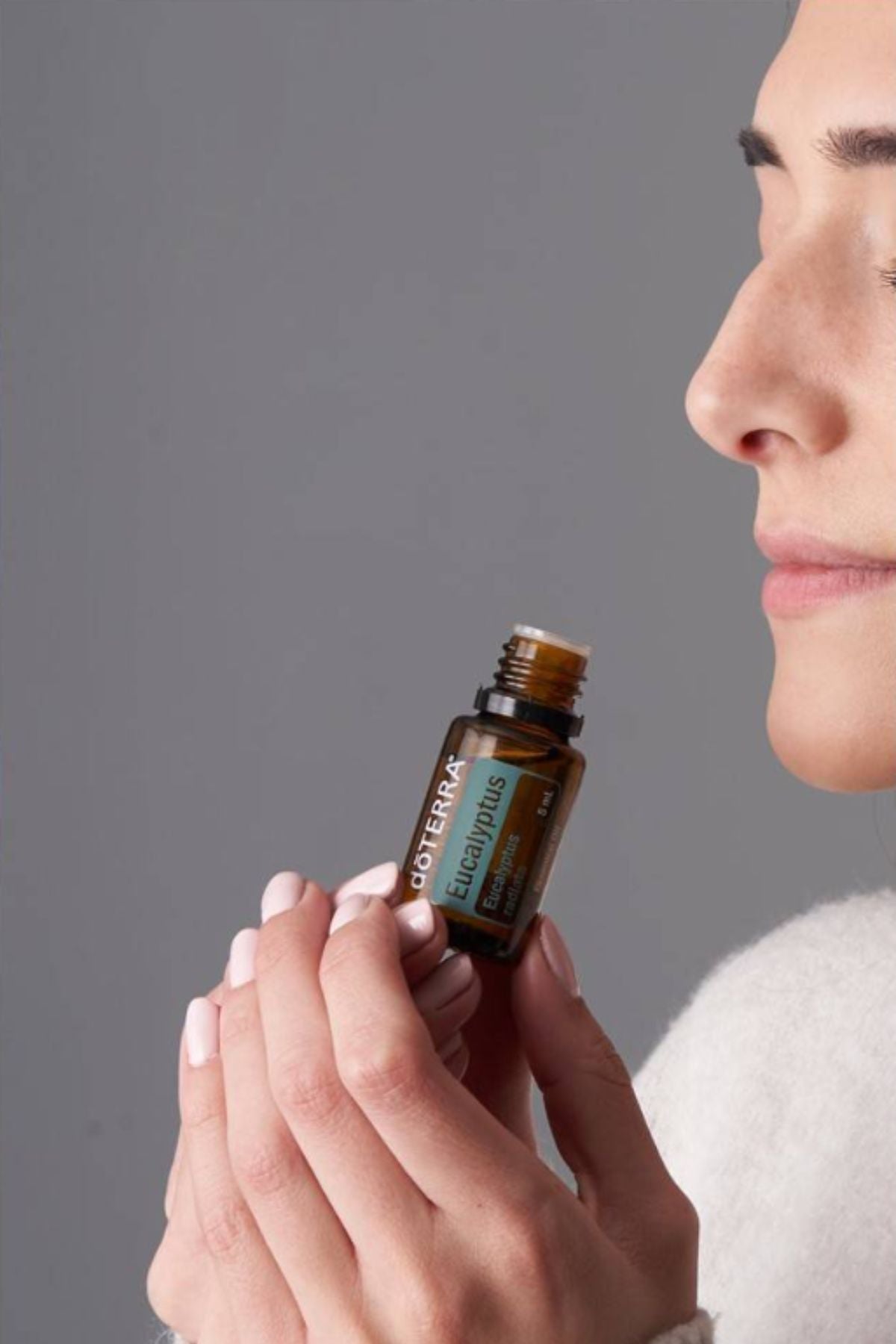 A close-up lifestyle shot of a woman holding a small amber glass bottle of doTERRA Eucalyptus essential oil, with her face partially visible in profile against a solid gray background.