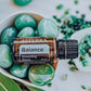 A bottle of doTERRA Balance Grounding Blend essential oil surrounded by green stones and leaves in a styled flat-lay arrangement.