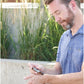 A man in a blue polo shirt outdoors holds a small amber bottle in his hands, looking down at it. Tall green reeds and a concrete planter are blurred in the background.