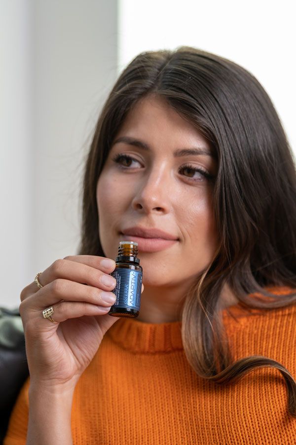 A woman with long brown hair holds a small amber glass essential oil bottle near her face. She is wearing an orange knit sweater, and the bottle label appears to read 'PURA' on a white background.