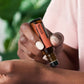 A close-up of a person holding a Neroli essential oil roller bottle over their wrist, with green leaves and a soft pink garment blurred in the background.