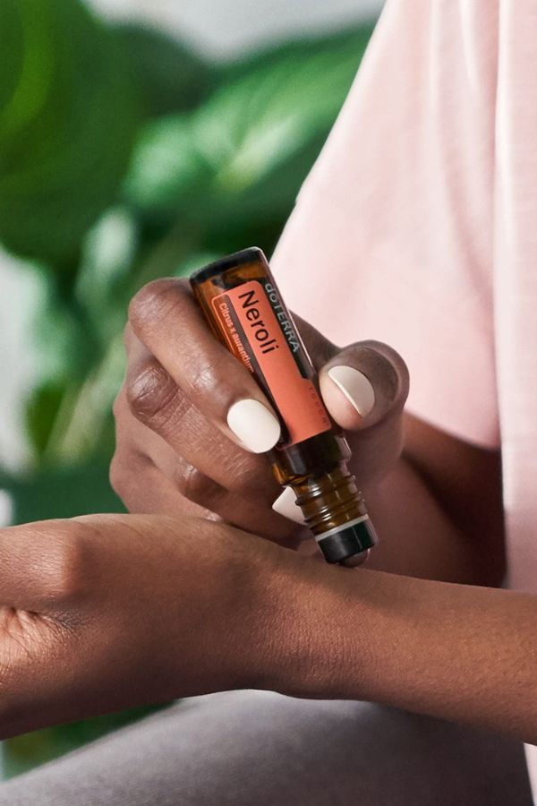 A close-up of a person holding a Neroli essential oil roller bottle over their wrist, with green leaves and a soft pink garment blurred in the background.