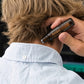 A person with light brown hair seen from behind wearing a light blue button-down shirt, while another hand holds an Osmira Amavi essential oil rollerball bottle near the base of the neck. doTERRA Amavi Touch Roll-on