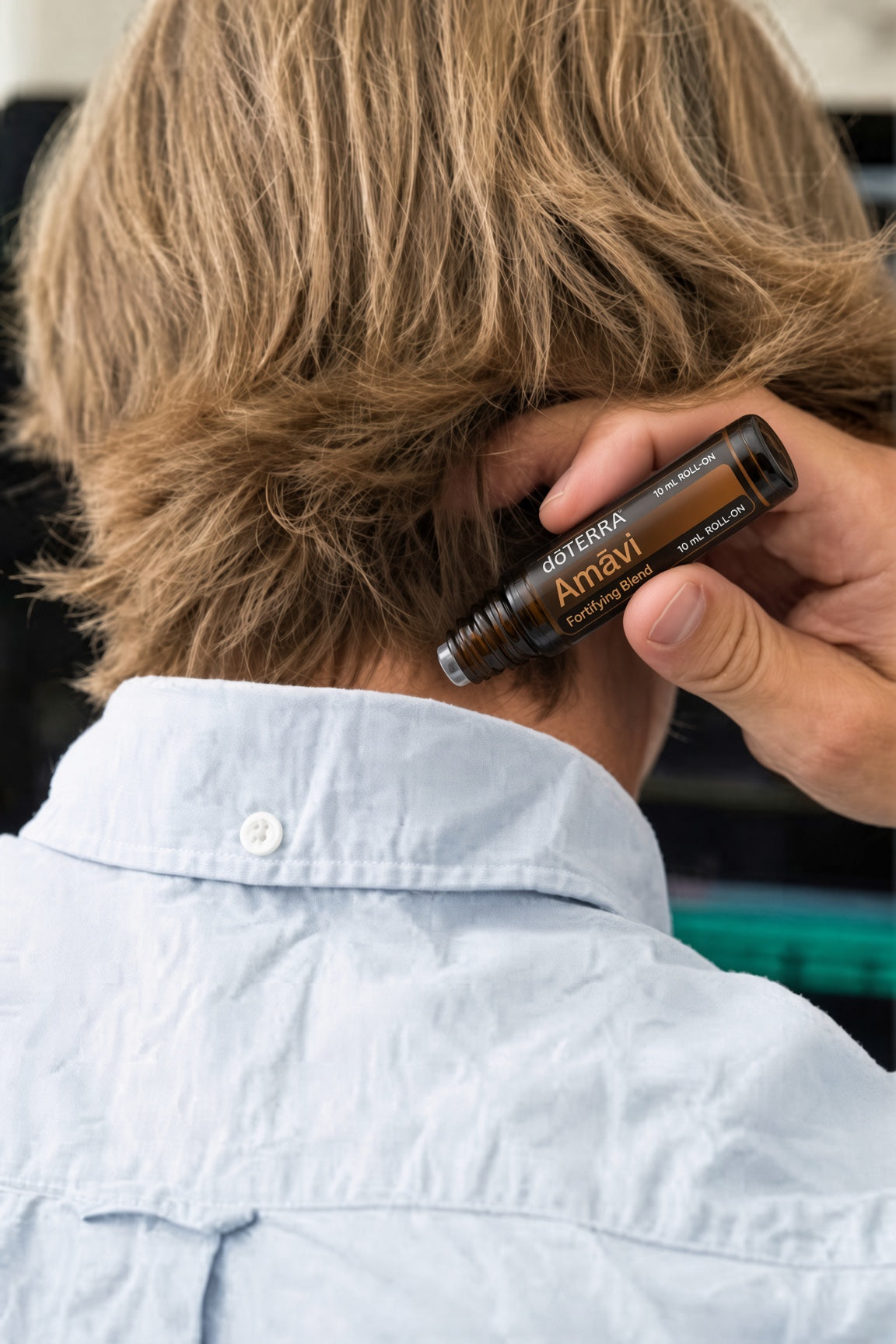 A person with light brown hair seen from behind wearing a light blue button-down shirt, while another hand holds an Osmira Amavi essential oil rollerball bottle near the base of the neck. doTERRA Amavi Touch Roll-on