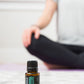 A small doTERRA Balance essential oil bottle sits on a light textured rug in the foreground, with a person in a gray shirt and black pants sitting cross-legged in a softly blurred indoor setting in the background.