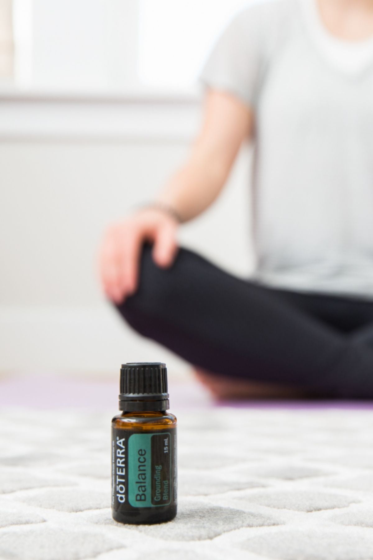 A small doTERRA Balance essential oil bottle sits on a light textured rug in the foreground, with a person in a gray shirt and black pants sitting cross-legged in a softly blurred indoor setting in the background.