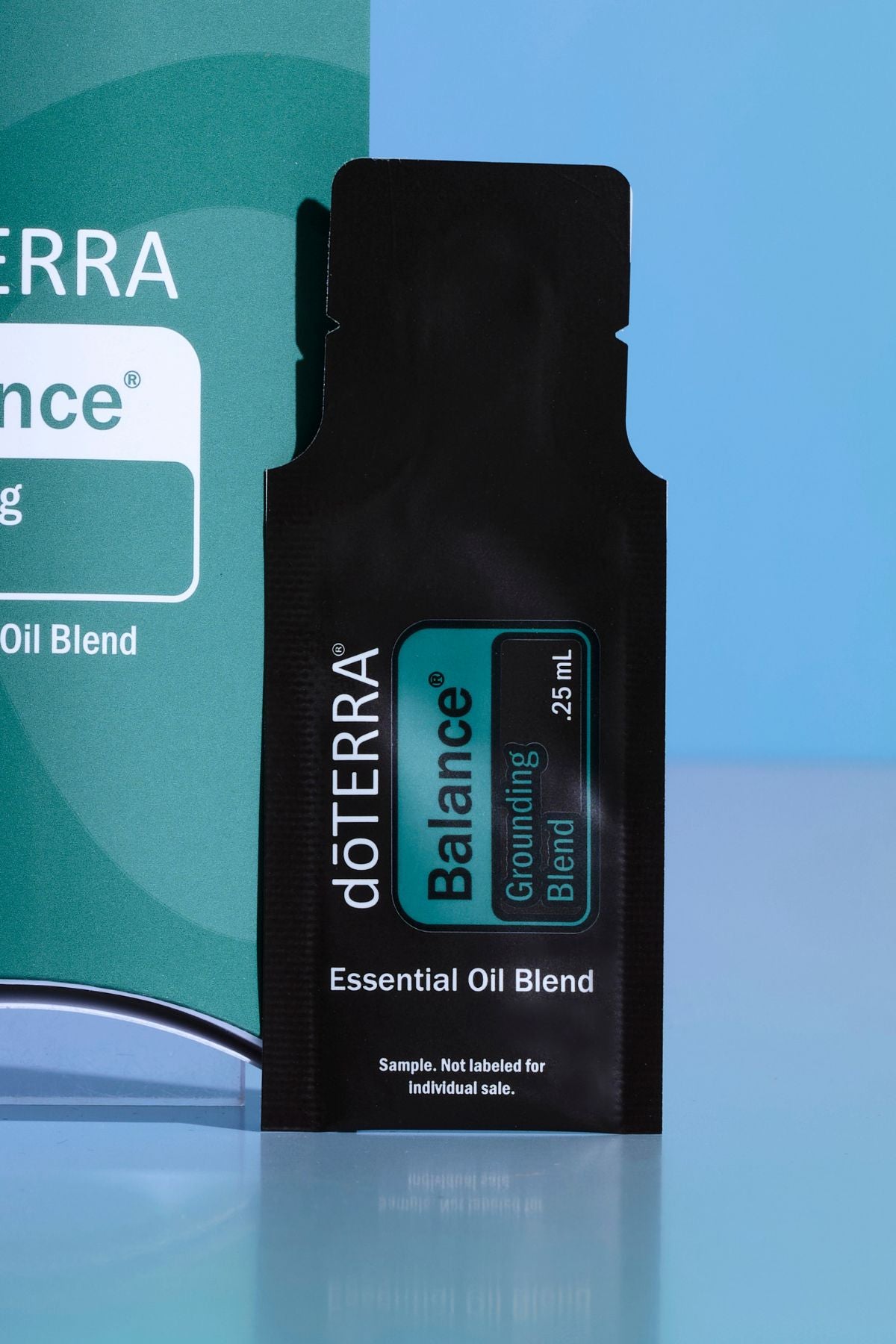 A single doTERRA Balance Grounding Blend essential oil blend sample sachet in black packaging, standing upright in front of a teal and blue branded box on a reflective light blue surface. doTERRA Balance Essential Oil Sample (.25 mL)