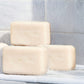 Three bars of Terra natural soap stacked on a white marble ledge under a shower stream, with a gray tiled wall in the background.