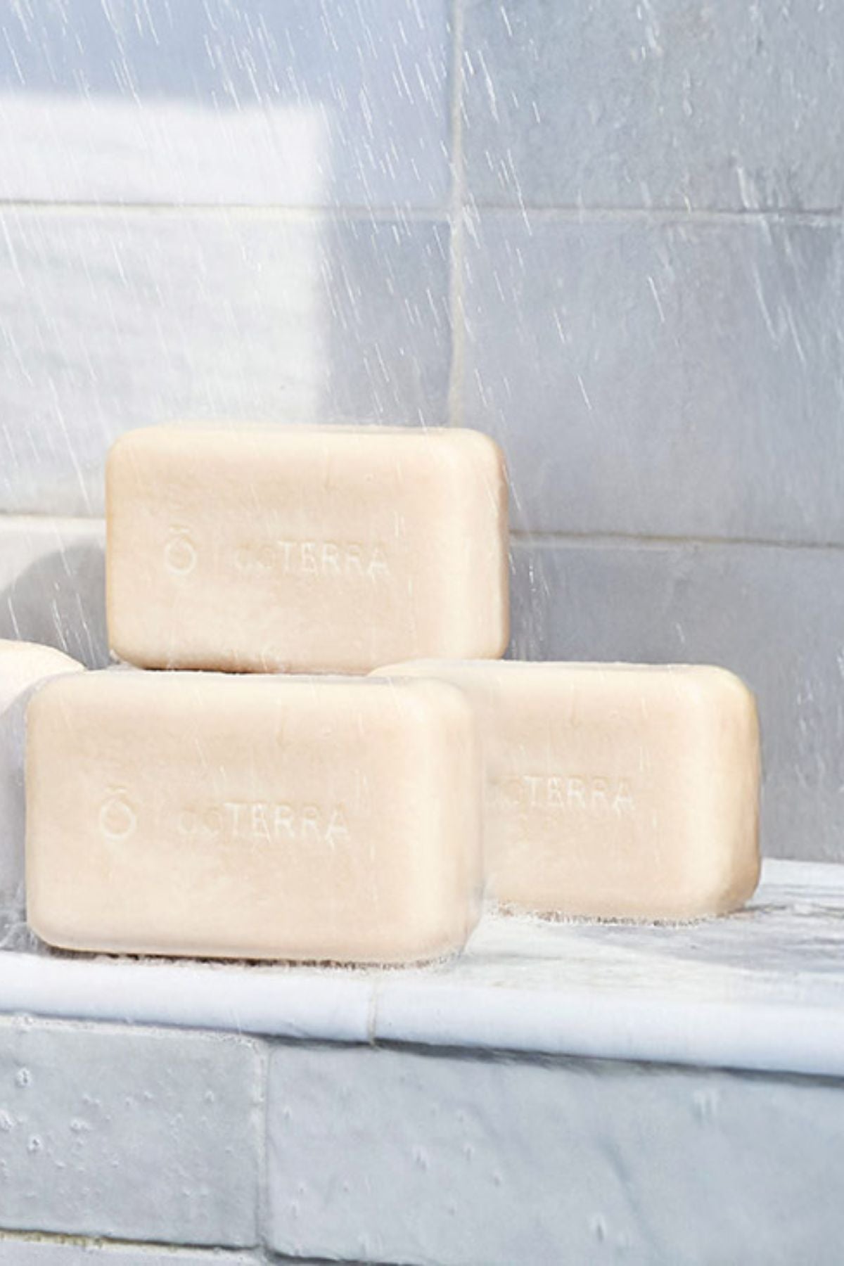 Three bars of Terra natural soap stacked on a white marble ledge under a shower stream, with a gray tiled wall in the background.