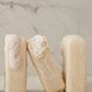 Three pale beige rectangular bars of soap standing on a light marble surface with a matching marble background. One soap is upright on the left with white lather on top, one is leaning in the center with foam on top, and one is upright on the right.